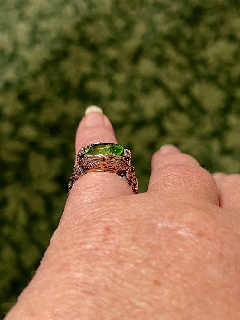May include: A silver ring with a green gemstone set in a detailed, intricate design. The ring is worn on a finger.
