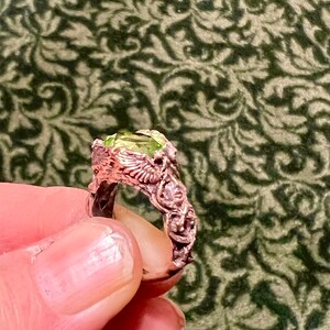 May include: A ring with a green gemstone set in a detailed, ornate, copper-colored band. The band is shaped like a pair of wings.