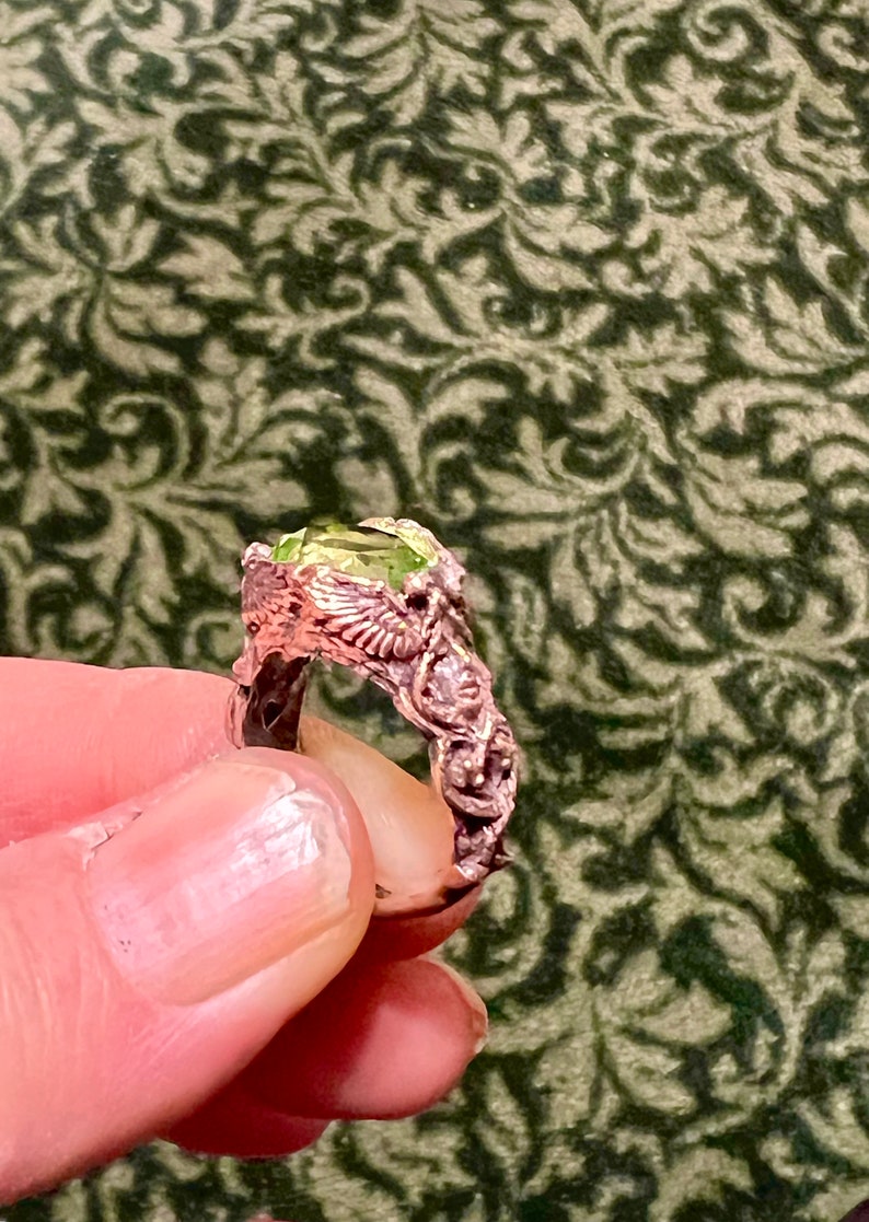 May include: A ring with a green gemstone set in a detailed, ornate, copper-colored band. The band is shaped like a pair of wings.