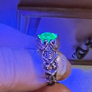 May include: A silver ring with a green gemstone set in a detailed, ornate design. The ring is on a hand, and the gemstone is glowing under a black light.