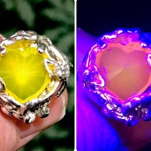 May include: A heart-shaped yellow gemstone ring set in a silver band with a unique, intricate design. The ring glows a bright pink under a UV light.