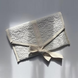 May include: An off-white quilted clutch bag with a textured pattern and a matching ribbon tie. The rectangular bag has a flap closure and a ribbon tied in a bow. The design is soft and neutral, suitable for various occasions.