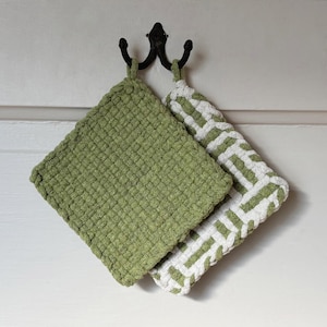 Puede incluir: Dos agarraderas de olla tejidas en verde y blanco colgadas de un gancho. Las agarraderas están hechas de una tela texturizada y tienen un diseño simple.