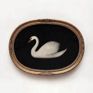 Peut inclure: Peinture encadrée ovale représentant un cygne blanc sur fond noir. Le cygne a un cou courbé et des plumes d'ailes détaillées. Le cadre doré présente des détails décoratifs, créant une esthétique classique.