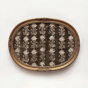 Peut inclure: Un plateau décoratif de forme ovale doté d'un cadre en métal doré aux détails ornés. La surface intérieure est recouverte d'un tissu marron foncé présentant un motif vertical répétitif de fleurs claires et de fines rayures.