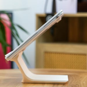 May include: A white tablet propped up on a matching stand, angled for viewing. The stand has a curved design, and the tablet is positioned on a wooden table. The background includes a microphone and a cabinet.
