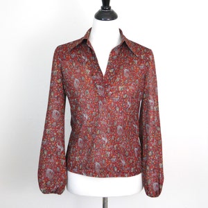 Può includere: Una blusa rossa a maniche lunghe con una stampa floreale con piccoli dettagli bianchi e dorati. La blusa ha un colletto e una chiusura con bottoni sul davanti.