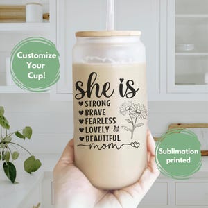 K&ouml;nnte beinhalten: Ein durchsichtiger Glasbecher mit braunem Deckel und Strohhalm. Der Becher hat einen wei&szlig;en Hintergrund mit schwarzem Text, der "She is Strong Brave Fearless Lovely Beautiful Mom" lautet, mit einem kleinen Schmetterling und einem Blumenmotiv.