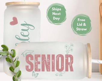 May include: A frosted glass tumbler with a bamboo lid and straw. The tumbler has a pink and green design with the word "SENIOR" in pink with a distressed look. The tumbler also has a graduation cap, butterfly, and diploma illustrations.