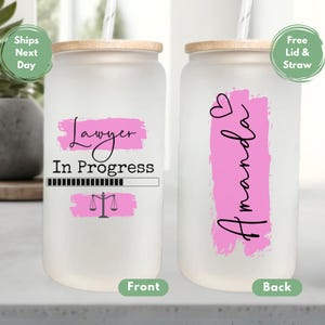 May include: A white tumbler with a pink and black design. The front of the tumbler says "Lawyer In Progress" with a progress bar and a scale of justice. The back of the tumbler says "Amanda" with a pink heart.