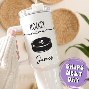 May include: White tumbler with a handle, featuring the text "HOCKEY mama" above a black hockey puck graphic with the number 6. Below the puck is the name "James" in cursive. The tumbler has a clear lid and a straw.
