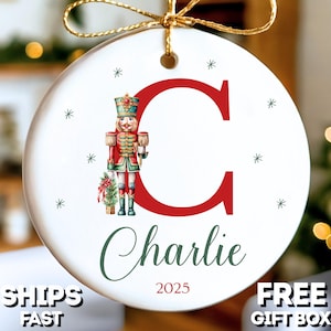 May include: White ceramic Christmas ornament with a red letter "C" and a nutcracker illustration. The ornament features the name "Charlie" and the year "2025". It is decorated with small snowflakes and a gold string for hanging.