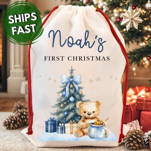 May include: A white drawstring Christmas gift bag with red cords, featuring the text "Noah's FIRST CHRISTMAS". The bag has a Christmas tree, teddy bear, and presents illustration. A green circle with "SHIPS FAST" is in the upper left.