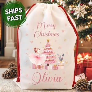 May include: A white drawstring Christmas gift bag with a red cord. The bag features a pink ballerina, Christmas tree, and the words "Merry Christmas" and "Olivia" in pink. The bag has a green "SHIPS FAST" sticker.