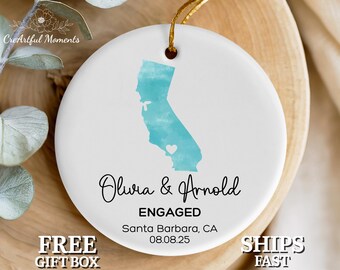 May include: White ceramic ornament with a teal watercolor depiction of the California state outline, featuring a heart. The ornament is personalized with the names "Olivia & Arnold" and the text "ENGAGED" and "Santa Barbara, CA 08.08.25".