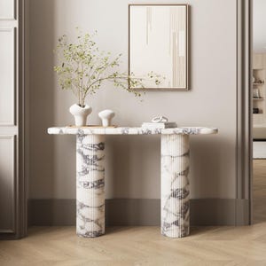 Puede incluir: Una consola con tapa de mármol y patas cilíndricas estriadas. La mesa presenta un patrón veteado blanco y gris. Una obra de arte enmarcada cuelga sobre la mesa, y jarrones y objetos decorativos se exhiben sobre la encimera. La habitación tiene paredes de color claro y suelo de madera.