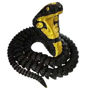 May include: A coiled articulated cobra figurine featuring a textured black body with a striking metallic gold hood and underbelly. The snake is depicted in an upright, alert posture with its hood flared, showcasing a high-contrast color scheme.