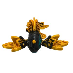 May include: A 3D-printed sculpture of a stylized insect with a black body, yellow wings, and a yellow head crest. The wings have a patterned design. The insect has teal accents on its face and a yellow dot on its nose.