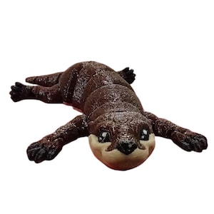May include: A brown otter figurine with outstretched paws and a light beige face. The otter's fur has a textured appearance, and its eyes are black. The figurine is lying down on a white surface.
