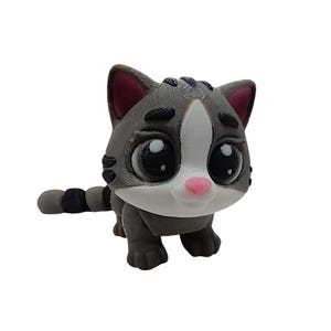May include: A gray and white cat figurine with large, expressive eyes and a pink nose. The cat has a striped tail and is posed in a standing position. The figurine is likely a decorative item or toy.