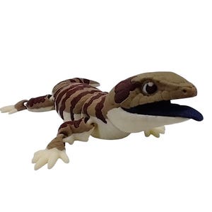 Peut inclure: Une peluche représentant un lézard à langue bleue. Le corps est rayé de beige et de marron, la tête est beige avec des taches marron foncé et la langue est bleue. Les pattes sont beige clair.