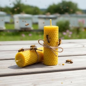 Puede incluir: Dos velas amarillas de cera de abeja con un patrón de panal, atadas con hilo de bramante y rodeadas de abejas. Las velas son cilíndricas y están sobre una superficie de madera, con un fondo borroso de colmenas y vegetación.