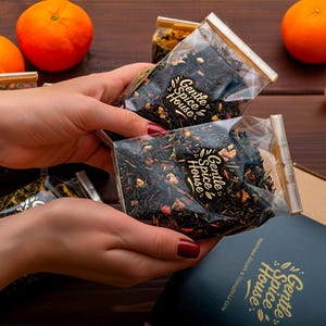 Op de afbeelding: Twee transparante zakjes gevuld met losse thee, vastgehouden door handen met rode nagellak. De zakjes zijn geseald met goud en hebben het opschrift "Gentle Spice House". Sinaasappels zijn zichtbaar op de achtergrond.