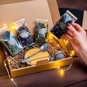 Op de afbeelding: Een geschenkdoos met thee, een lepel en een thee-ei. De doos bevat vier doorzichtige theezakjes met de tekst "Gentle Spice House". De doos is gevuld met bruin verpakkingsmateriaal en lichtslingers.