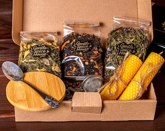 May include: A gift box containing tea, candles, and accessories. The box includes three sealed bags of tea, a wooden spoon, a tea infuser, and two yellow beeswax candles tied with twine. The bags are labeled "Gentle Spice House".
