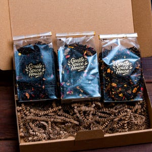 Op de afbeelding: Een kartonnen doos gevuld met drie doorzichtige theezakjes. Elk zakje heeft een label met de tekst "Gentle Spice House". De thee lijkt een mix van donkere bladeren en kleurrijke gedroogde bloemen. De doos bevat bruin crêpepapier.