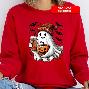 Puede incluir: Sudadera roja con un fantasma blanco que lleva una boina de color calabaza, sosteniendo una calabaza y una taza de bebida naranja. El fantasma est&aacute; rodeado de murci&eacute;lagos y estrellas.
