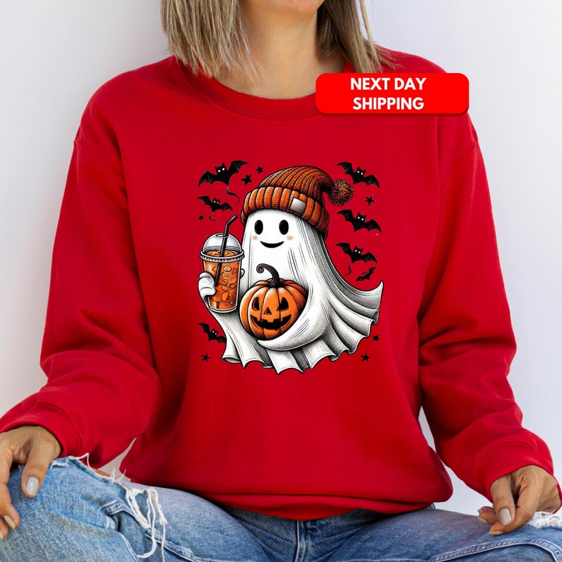 Puede incluir: Sudadera roja con un fantasma blanco que lleva una boina de color calabaza, sosteniendo una calabaza y una taza de bebida naranja. El fantasma est&aacute; rodeado de murci&eacute;lagos y estrellas.