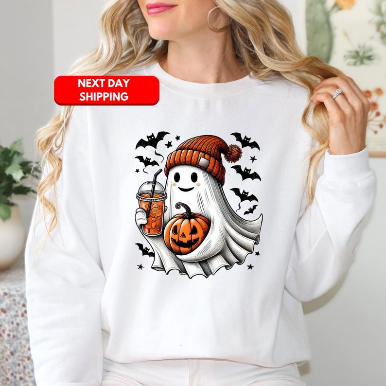 Puede incluir: Sudadera blanca con un lindo fantasma de dibujos animados que lleva un gorro y sostiene una calabaza y una taza de caf&eacute;. El fantasma est&aacute; rodeado de murci&eacute;lagos y estrellas. El texto "Next Day Shipping" est&aacute; en un cuadro rojo en la parte superior de la imagen.