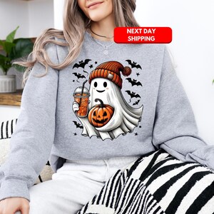 Puede incluir: Una sudadera gris con un lindo fantasma de dibujos animados sosteniendo una calabaza y una taza de caf&eacute;. El fantasma lleva un gorro rojo y blanco y tiene una cara amigable. La sudadera tiene un tema de Halloween con murci&eacute;lagos y estrellas.