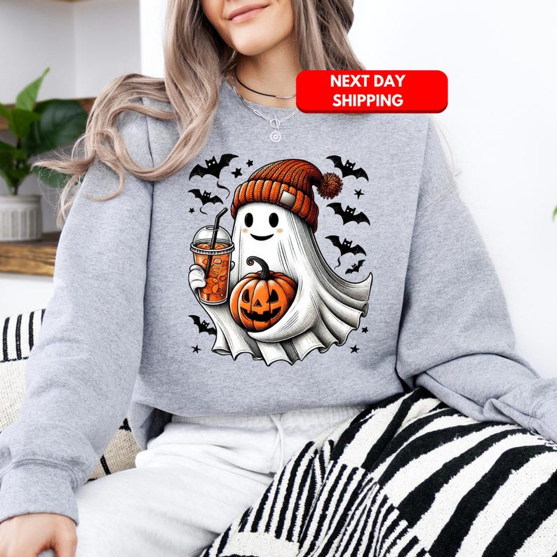 Puede incluir: Una sudadera gris con un lindo fantasma de dibujos animados sosteniendo una calabaza y una taza de caf&eacute;. El fantasma lleva un gorro rojo y blanco y tiene una cara amigable. La sudadera tiene un tema de Halloween con murci&eacute;lagos y estrellas.