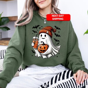 Puede incluir: Sudadera verde con un fantasma blanco que lleva un gorro de punto rojo y blanco, sosteniendo una calabaza y una taza de l&iacute;quido naranja. El fantasma tiene una cara amigable y est&aacute; rodeado de murci&eacute;lagos.