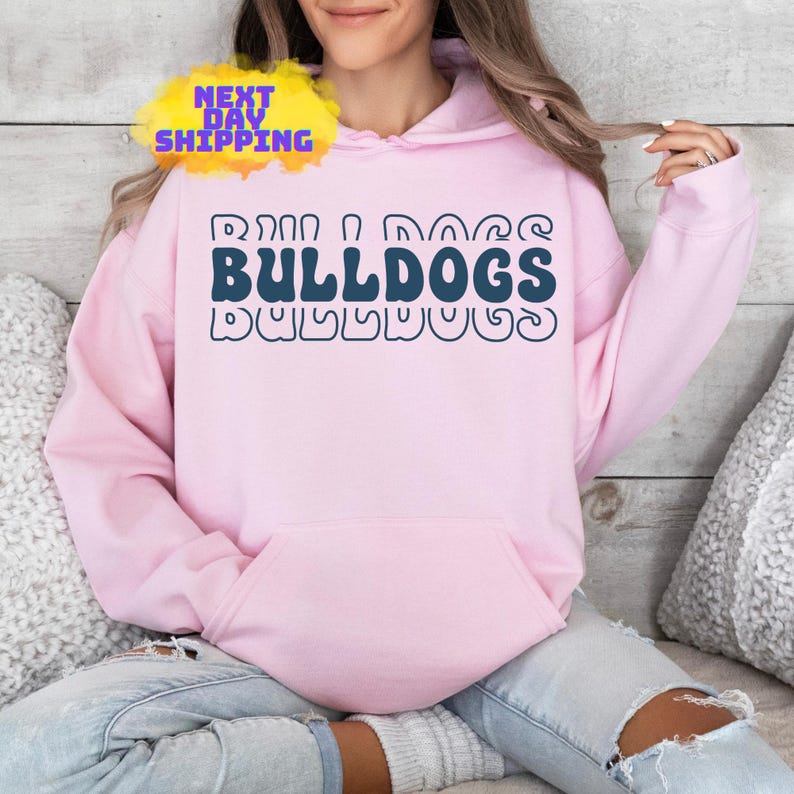 Op de afbeelding: Een roze hoodie met een blauwe en witte afbeelding met de tekst "Bulldogs" in een retro-lettertype.