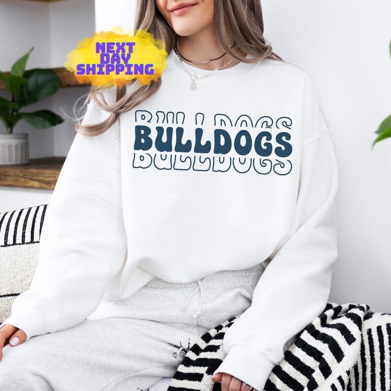 Op de afbeelding: Wit sweatshirt met een donkerblauwe grafische afbeelding met de tekst "Bulldogs" in een retro-lettertype.