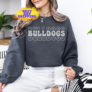 Op de afbeelding: Een donkergrijs sweatshirt met een witte afbeelding die "Bulldogs" zegt in een retro-lettertype.