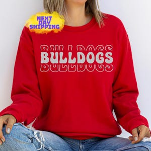 Op de afbeelding: Rood sweatshirt met de tekst "Run, Dog, Bulldogs" in een retro-lettertype.