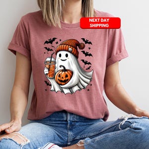 Puede incluir: Una camiseta rosa con un fantasma de dibujos animados que lleva un gorro y sostiene una calabaza y una taza de caf&eacute; helado. El fantasma est&aacute; rodeado de murci&eacute;lagos y estrellas.