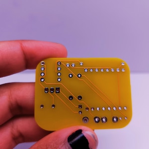Close-up of the back side of a yellow custom PCB held in a person's hand, showing clean copper traces and solder pads for through-hole components. The board is designed for an electronics DIY project.