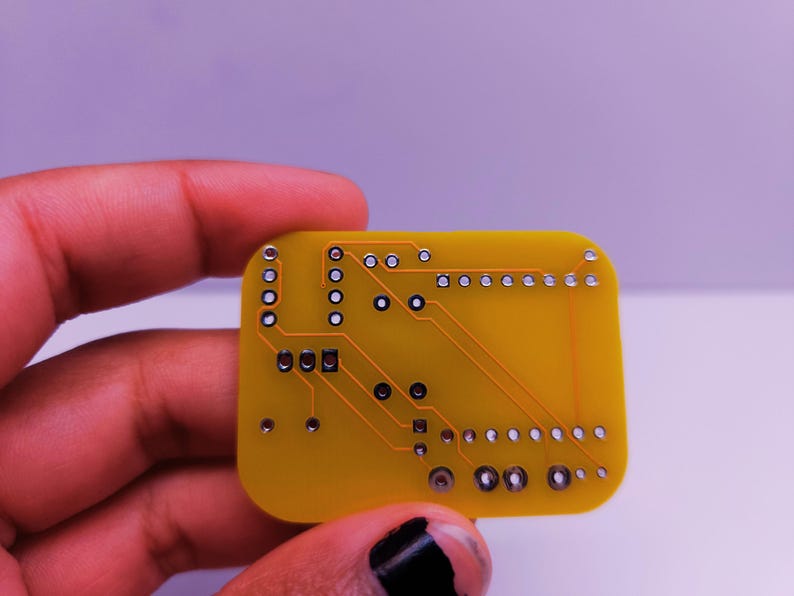Close-up of the back side of a yellow custom PCB held in a person's hand, showing clean copper traces and solder pads for through-hole components. The board is designed for an electronics DIY project.