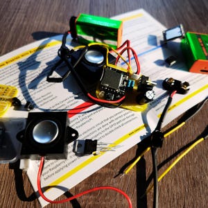 DIY electronics project laid out on top of a printed instruction manual on a wooden surface. Components include a pre-soldered PCB with a speaker and wires attached, a second bare PCB, microSD card, resistors, capacitors, 9V batteries, jumper wires, and other small parts.