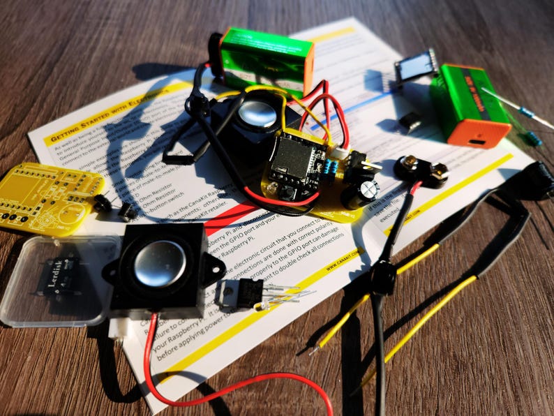 DIY electronics project laid out on top of a printed instruction manual on a wooden surface. Components include a pre-soldered PCB with a speaker and wires attached, a second bare PCB, microSD card, resistors, capacitors, 9V batteries, jumper wires, and other small parts.