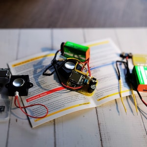 DIY electronics kit components spread across an open instruction manual on a wooden surface. Items include speakers, a pre-soldered circuit board, 9V batteries, jumper wires, a microSD card, and other electronic parts for assembly.