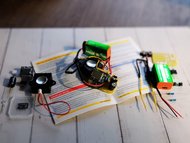 DIY electronics kit components spread across an open instruction manual on a wooden surface. Items include speakers, a pre-soldered circuit board, 9V batteries, jumper wires, a microSD card, and other electronic parts for assembly.