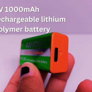 Close-up of a hand holding a green and orange ANVOW 9V 1000mAh rechargeable lithium polymer battery with a USB charging port. Overlaid text reads '9V 1000mAh rechargeable lithium polymer battery'