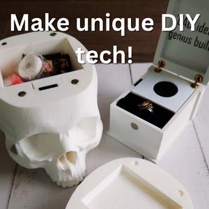 Photo of two 3D-printed DIY tech projects: a white skull-shaped container filled with candy and a small white music ring box displaying a ring. The lid of the ring box reads 'BAD ideas, genius built~'. Overlaid text says 'Make unique DIY tech!'
