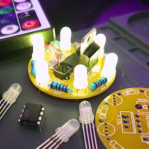 May include: A close-up of a yellow circuit board with several white LED lights illuminated. The board has resistors, a button, and other electronic components. Additional LEDs and components are scattered around the board. A remote control is visible in the background.